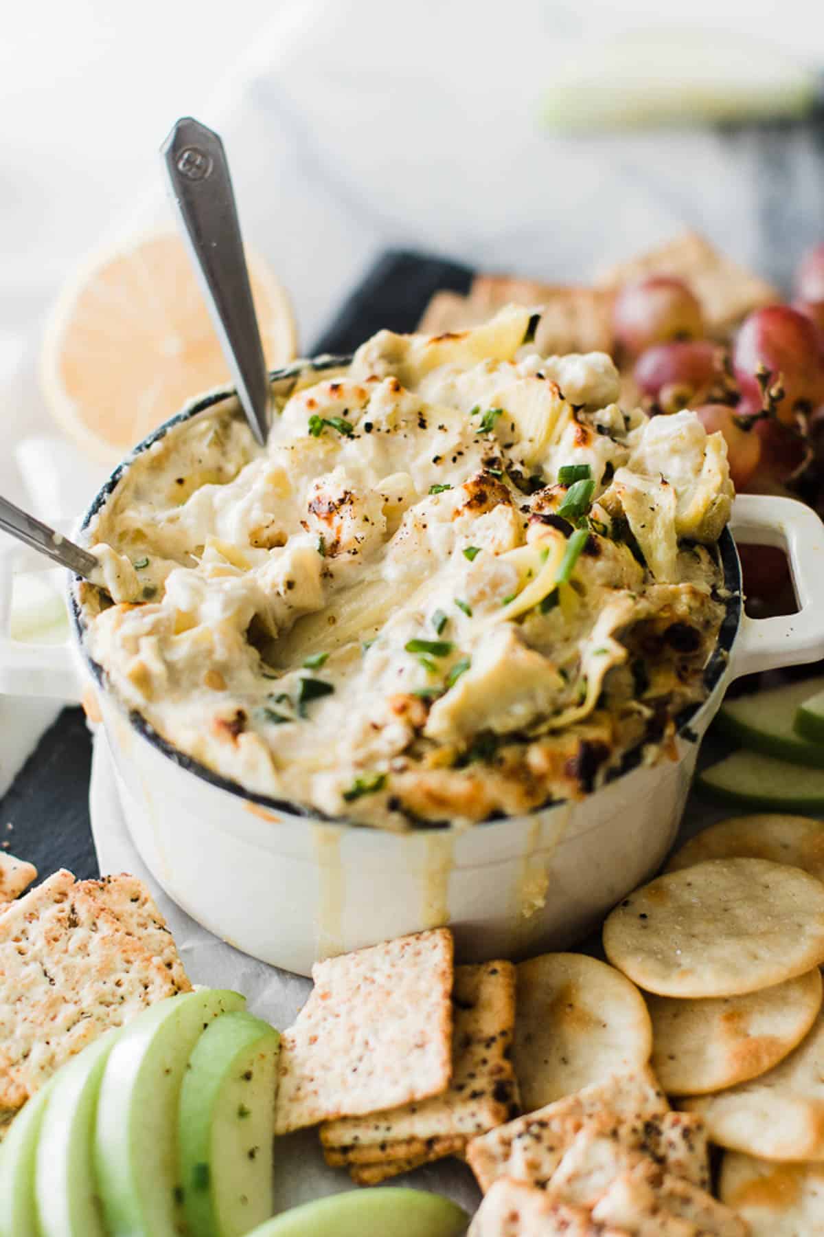 Bowl of green chile artichoke dip with spoons, crackers and fruit.