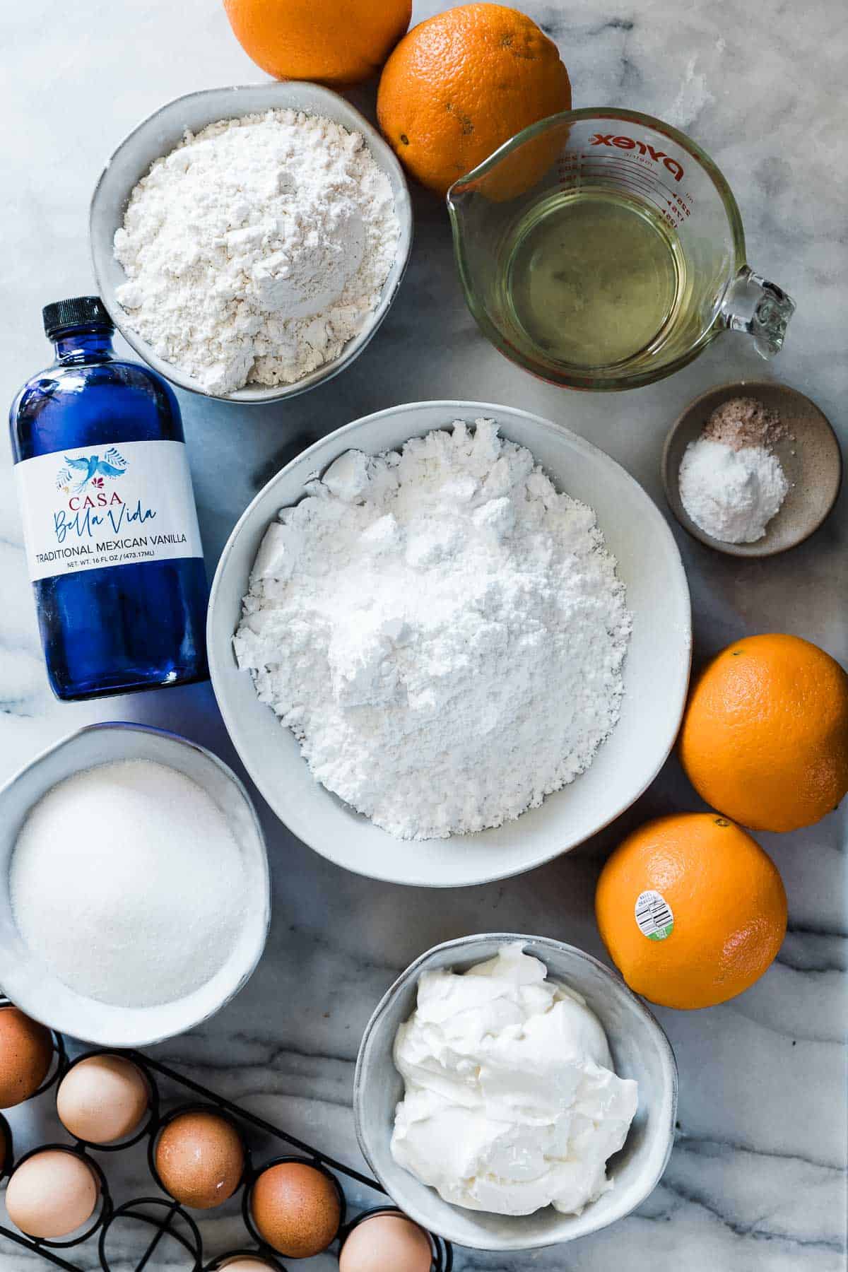 Flour, sugar, eggs, sour cream, and oranges on a marble counter.