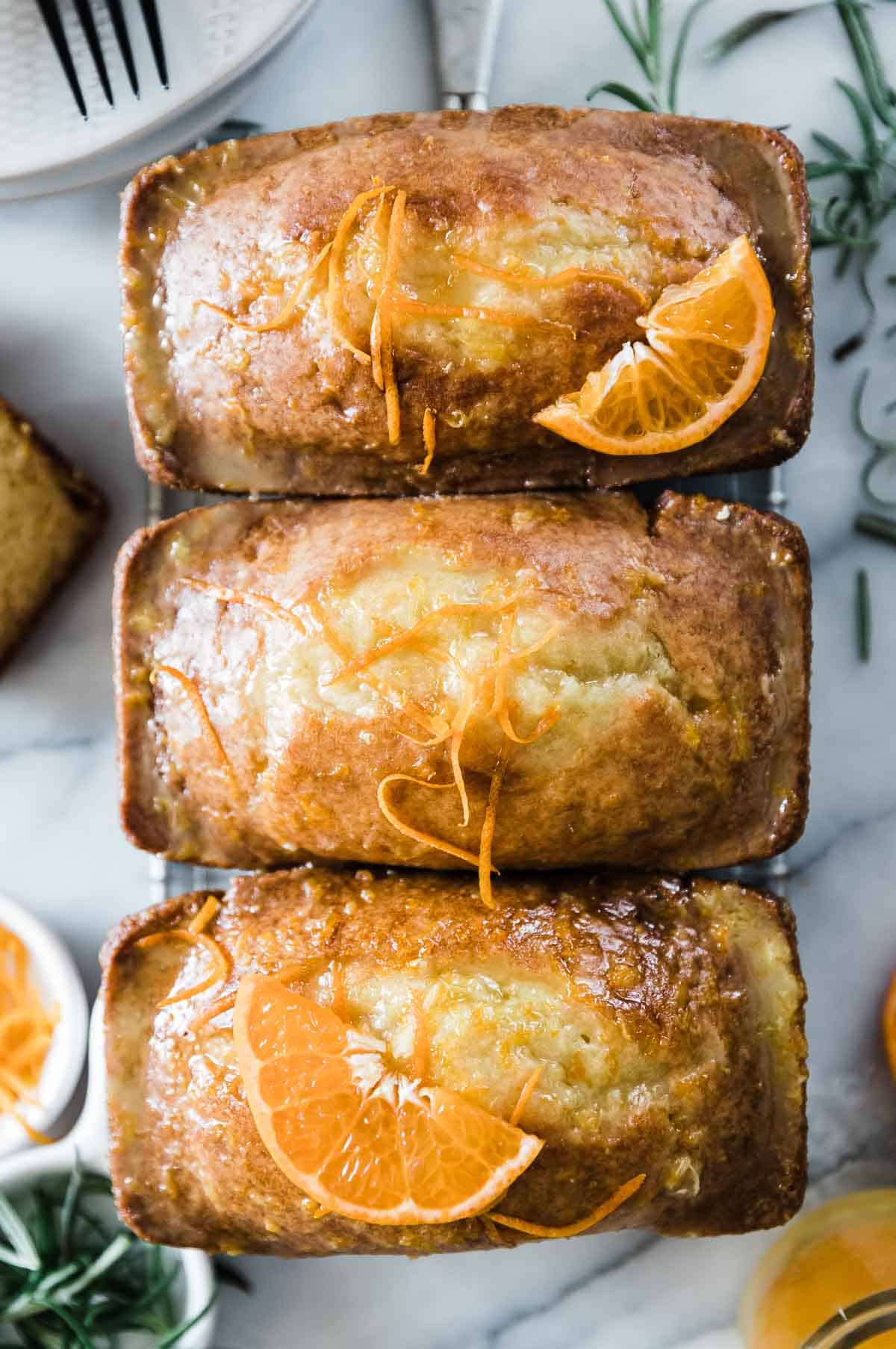Three loaves of orange bread on a wire cooling rack. They are garnished with orange zest.