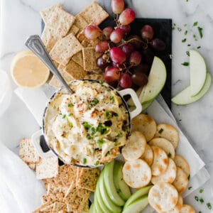 Hot artichoke dip surrounded by fruit, crackers and toasts on the table.