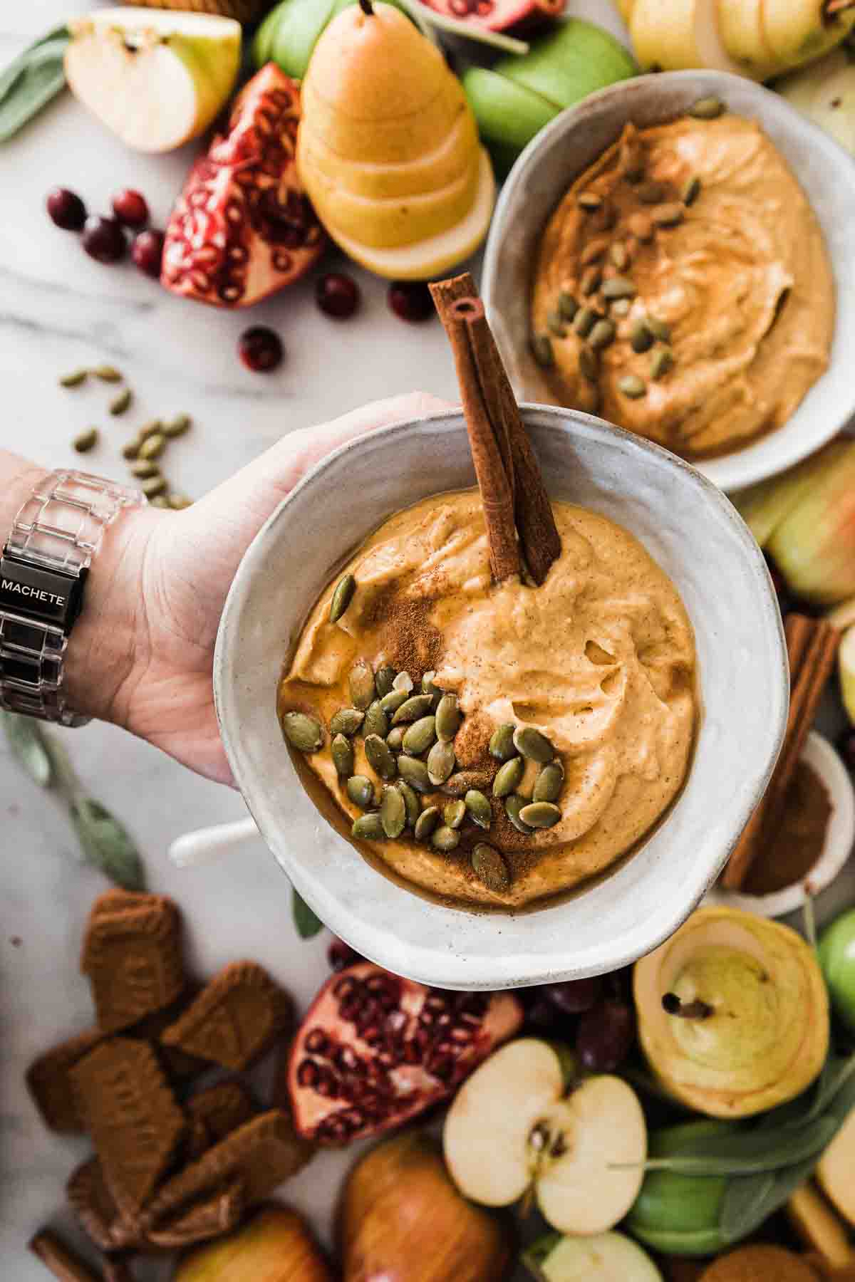 A grey bowl of pumpkin dip being held up. There is a cinnamon stick and pepitas garnishing the dip.