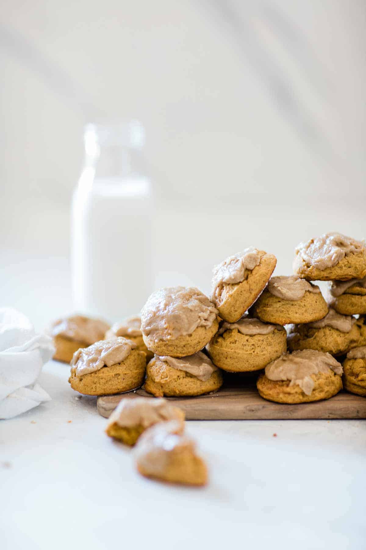 A large scatter of soft pumpkin cookies on a wooden board. There is a bottle of milk in the background.
