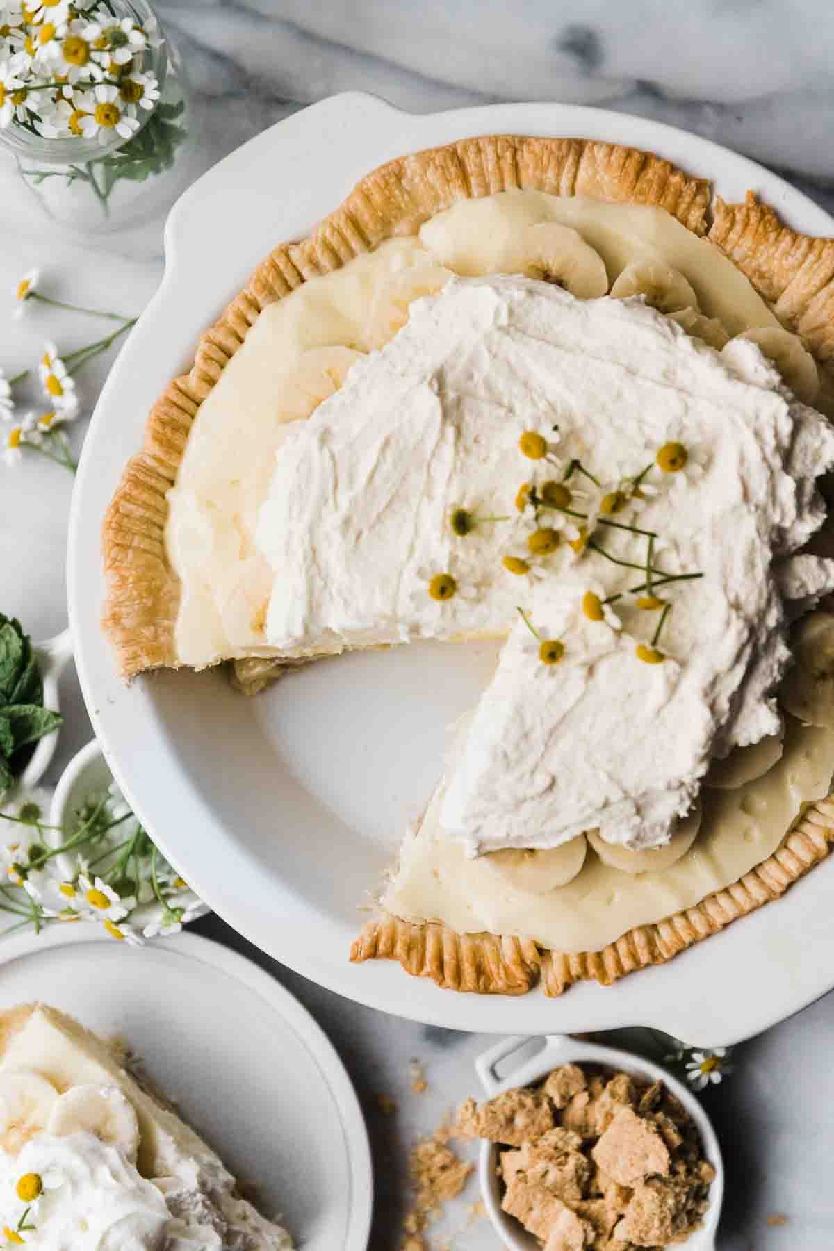 An overhead shot of easy banana cream pie in a white pie plate. It is garnished with whipped cream and chamomile flowers.