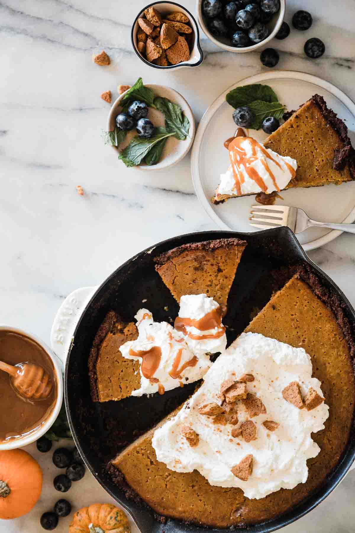 Easy pumpkin pie in a cast iron skillet. There are small bowls of blueberries, cookies, mint, and caramel to the side.