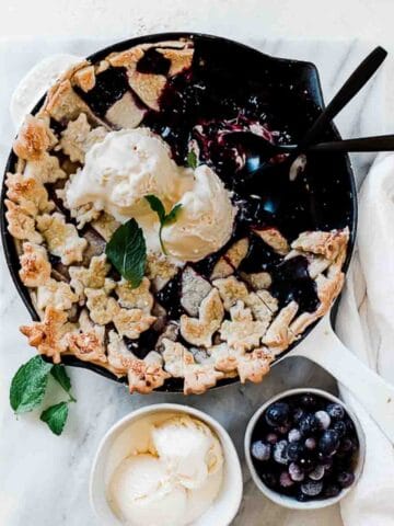razzleberry pie in a white cast iron pan on a marble counter. There is a bowl or berries and ice cream to the side.