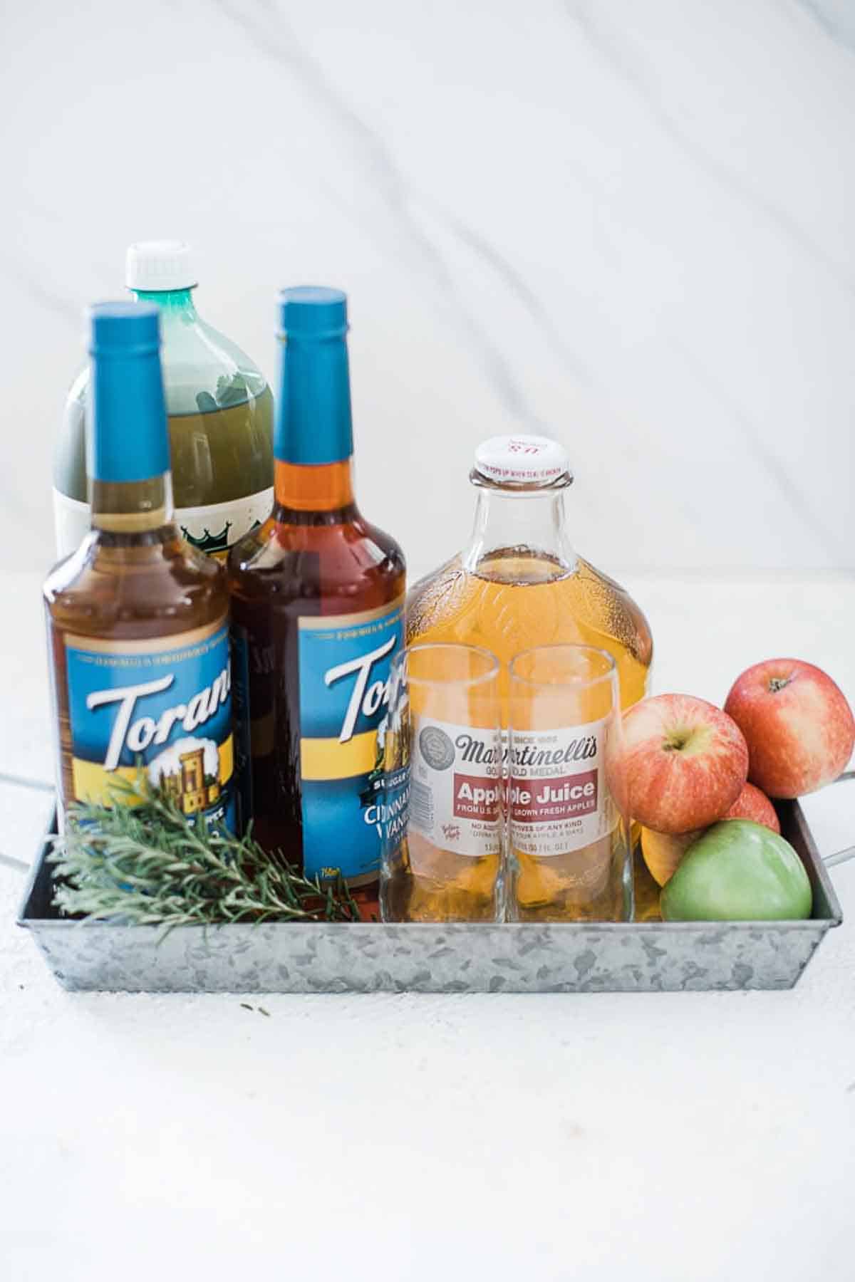 Flavored syrups, apple juice, ginger ale and apples in a metal tray.
