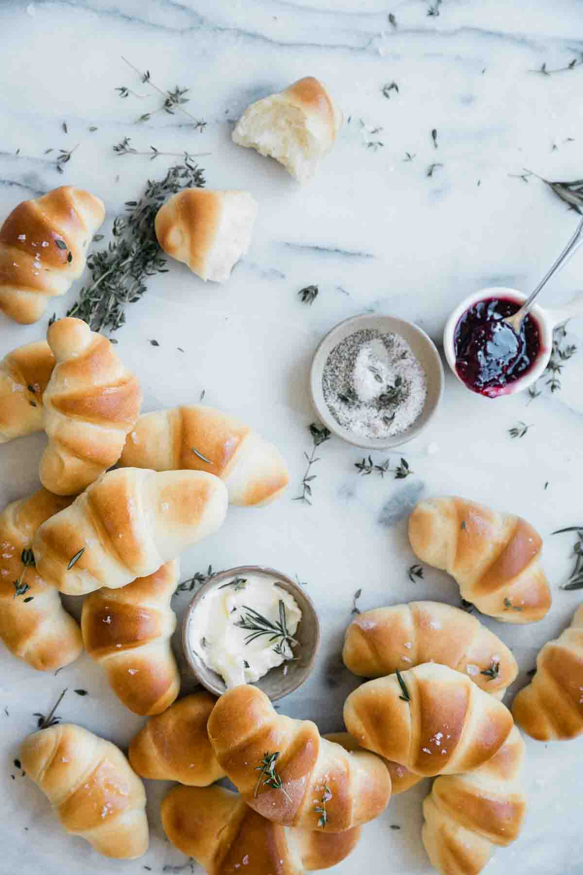 Crescent rolls scattered on a marble counter. There are small bowls of butter, jam, and salt and pepper to the side.