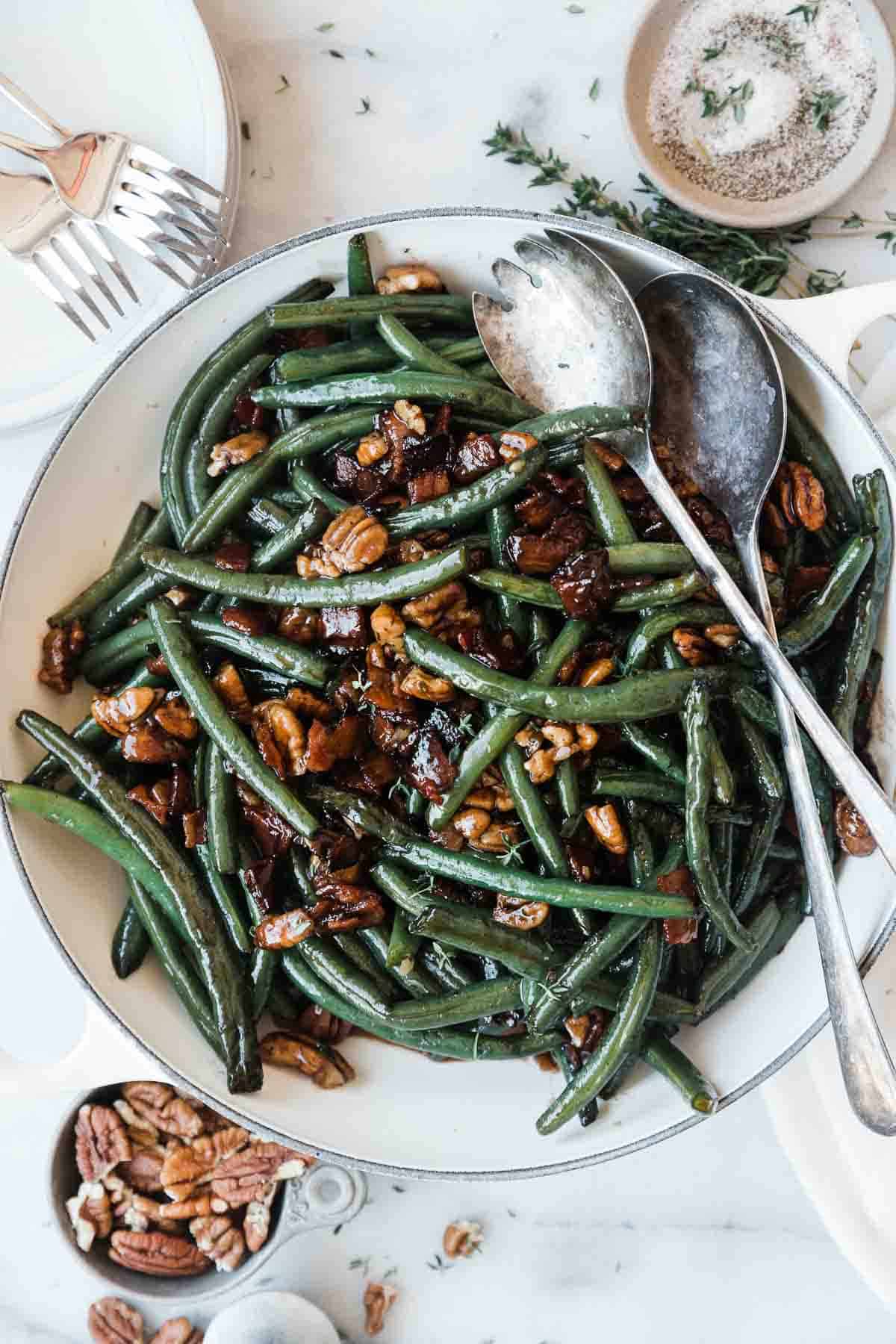 An overhead shot of green beans and bacon in a white braised. There is a small bowl of pecans on one corner and a small bowl of salt and pepper on the oven.