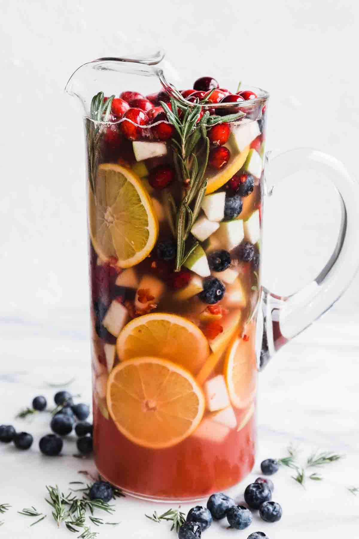 A tall pitcher filled with non alcoholic sangria. There is rosemary and blueberries to the side.
