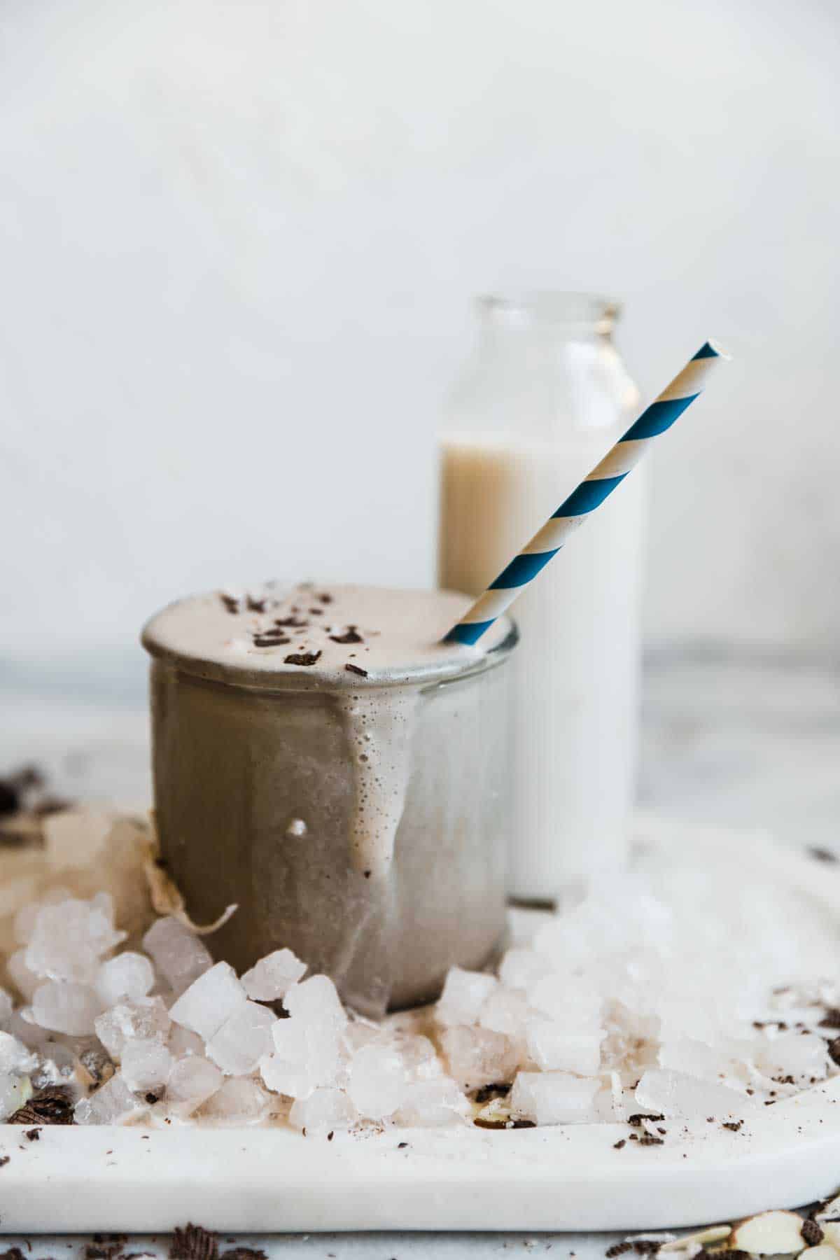 A single glass of chocolate protein smoothie with a straw. There is a bottle of milk in the background.