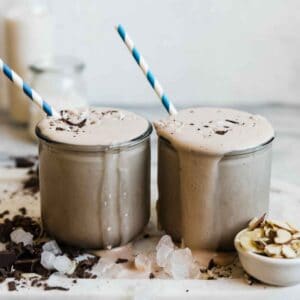 Two glasses filled with chocolate protein smoothie. They are surrounded by ice and shaved chocolate.