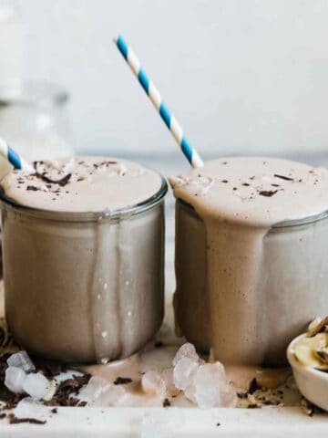 Two glasses filled with chocolate protein smoothie. They are surrounded by ice and shaved chocolate.