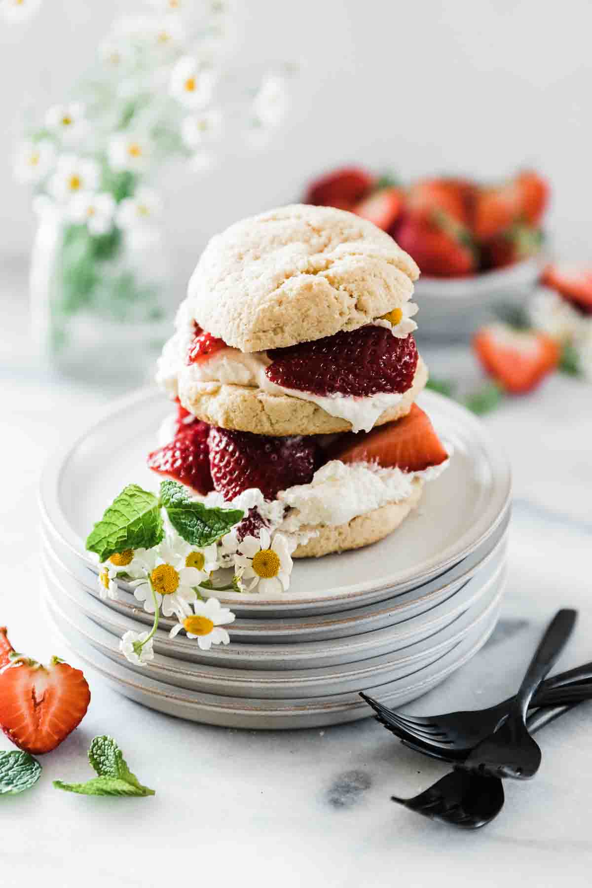 A single serve strawberry shortcake on a stack of grey dessert plates with flowers on the side.
