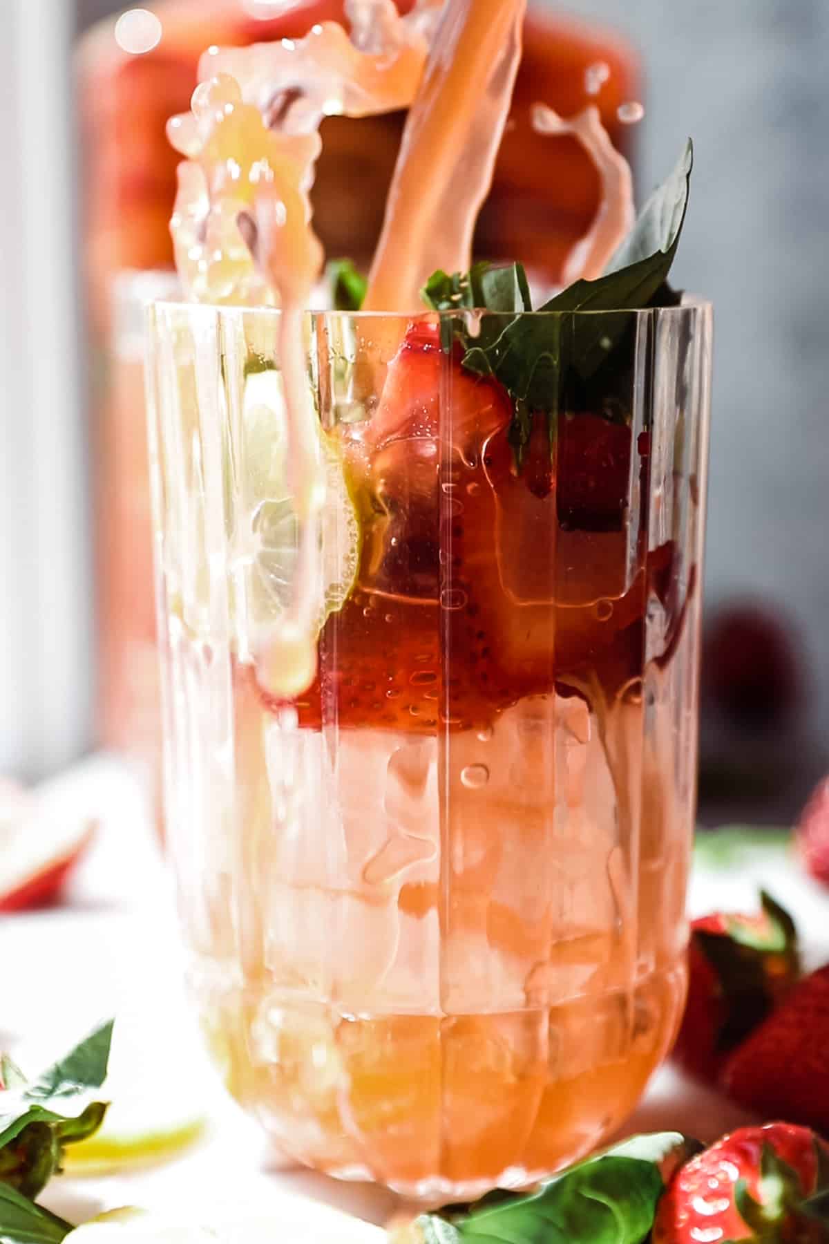 Large glass of ice with strawberry garnish with a splash of strawberry lemonade being poured in. 