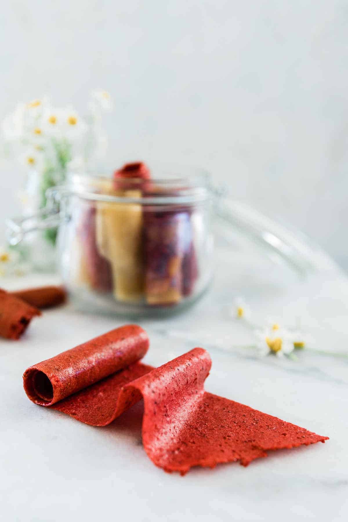 Homemade fruit leather slightly rolled out onto a marble counter. There is a container of fruit leathers in the background.