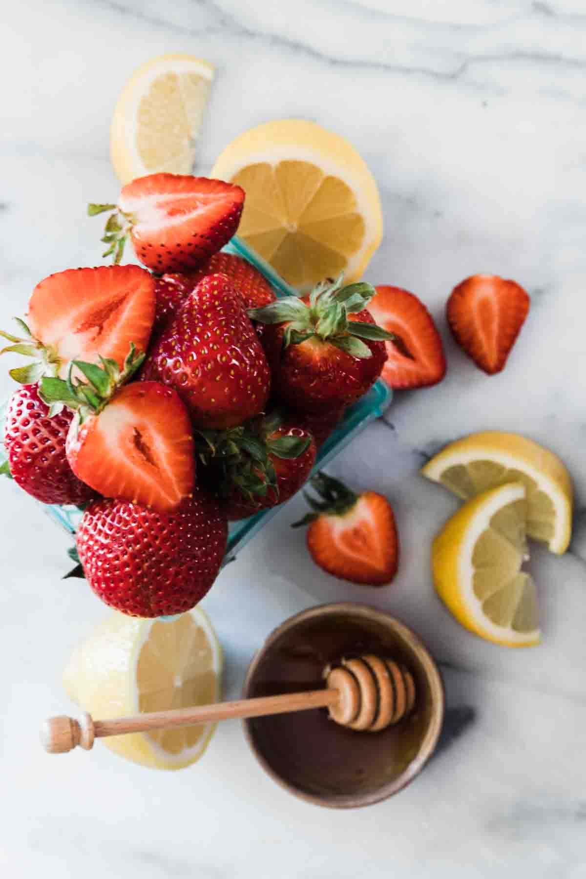 A pint of strawberries, some lemon slices, and a small bowl of honey on a marble counter.