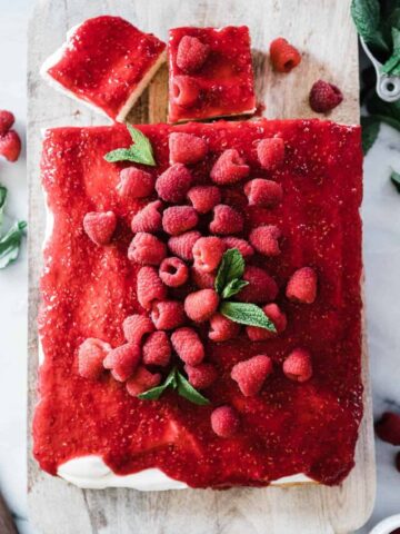 Raspberry cake recipe on a wooden cutting board. There are a few sliced cut out and it is garnished with raspberries.
