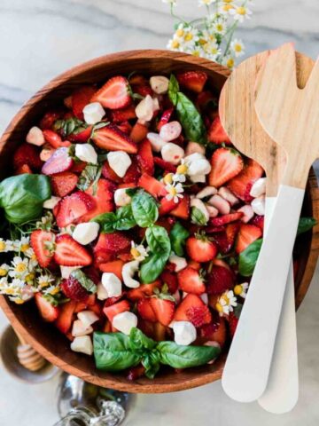 Strawberry caprese salad in a large wooden bowl. There are white salad servers to the side.