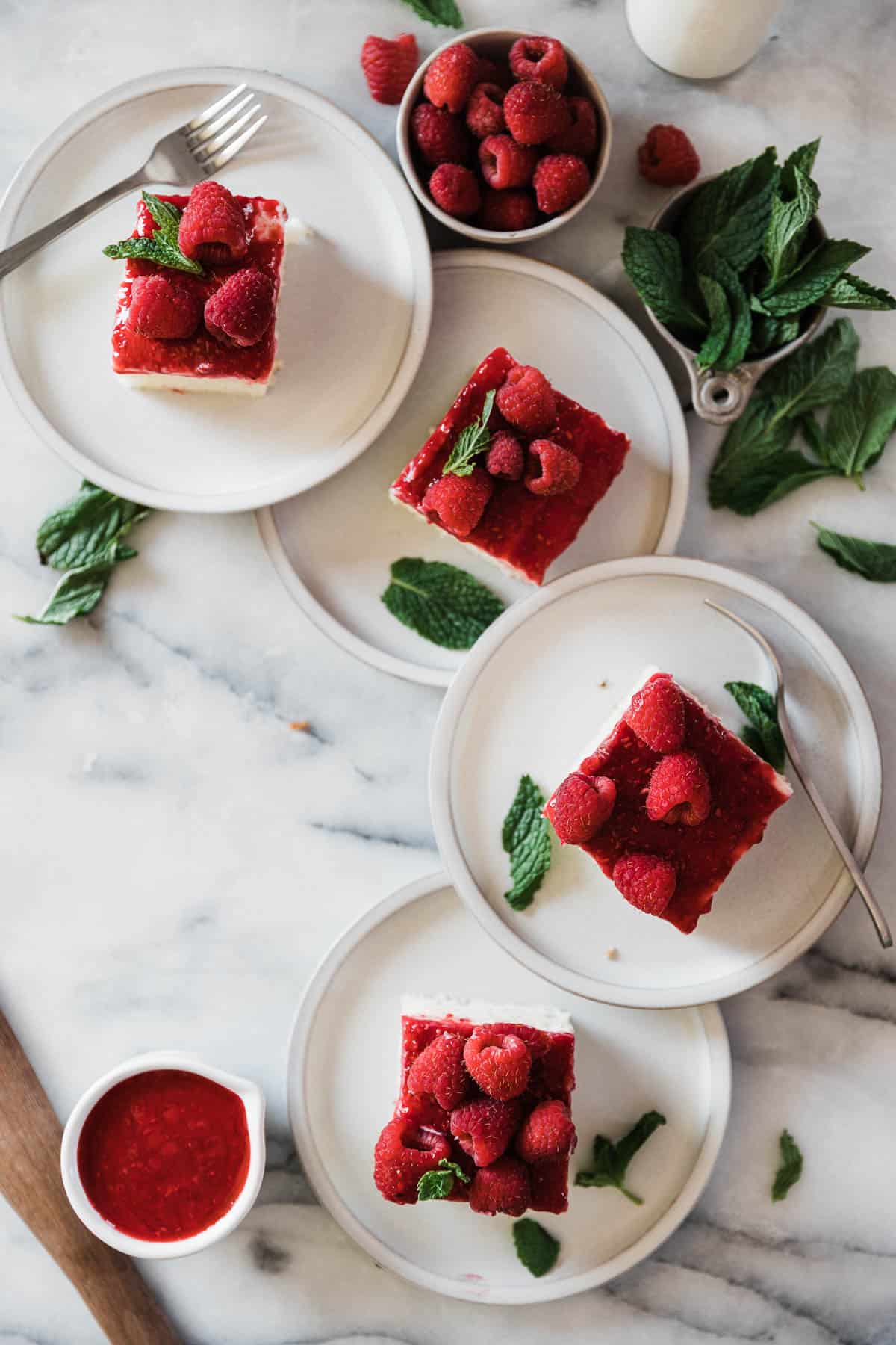 Four plates in a row, each with a slice of raspberry glazed cake. There is a small bowl of raspberries to the side and mint leave scattered.