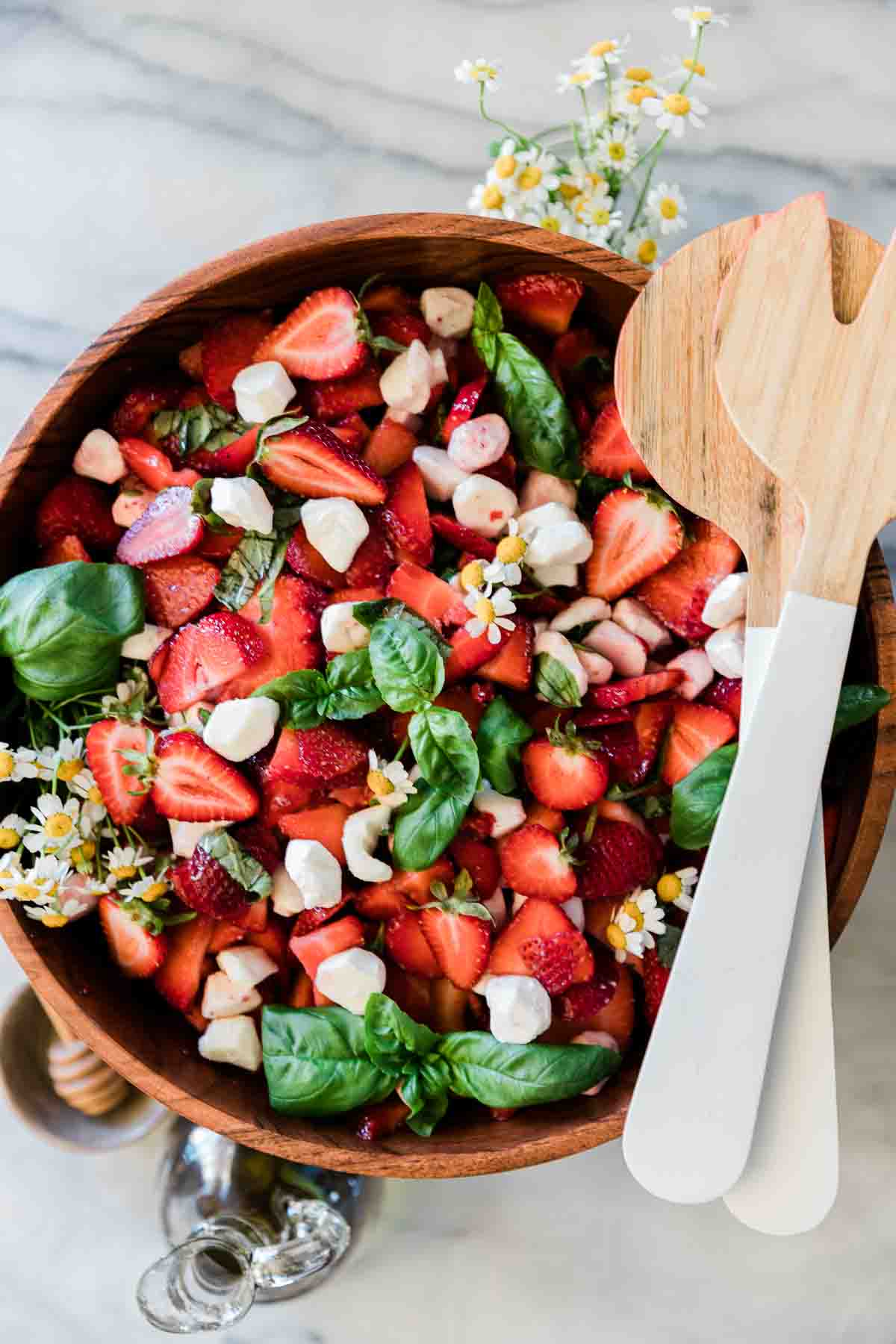 Strawberry caprese salad in a large wooden bowl. There are white salad servers to the side.
