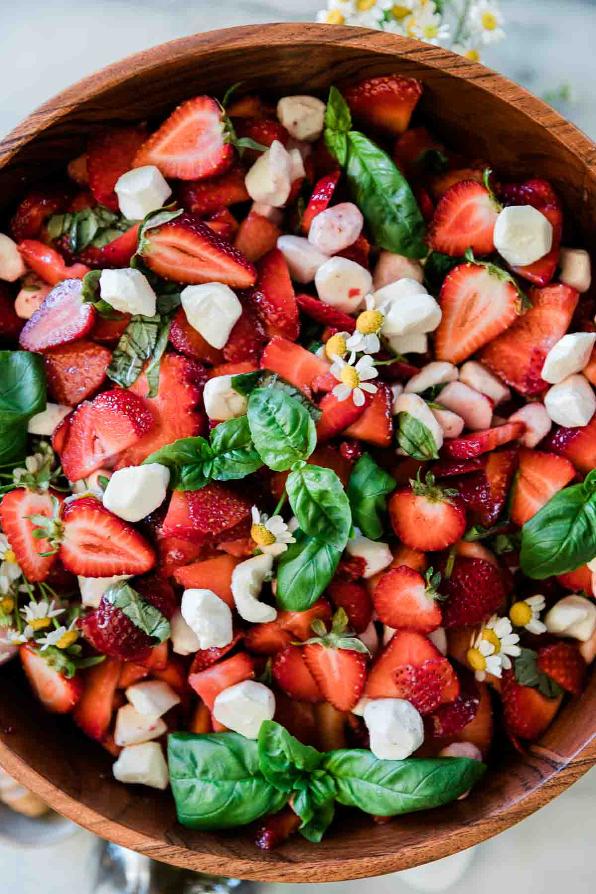 A close up of strawberry summer salad in a wooden bowl. It is garnished with basil and flowers.