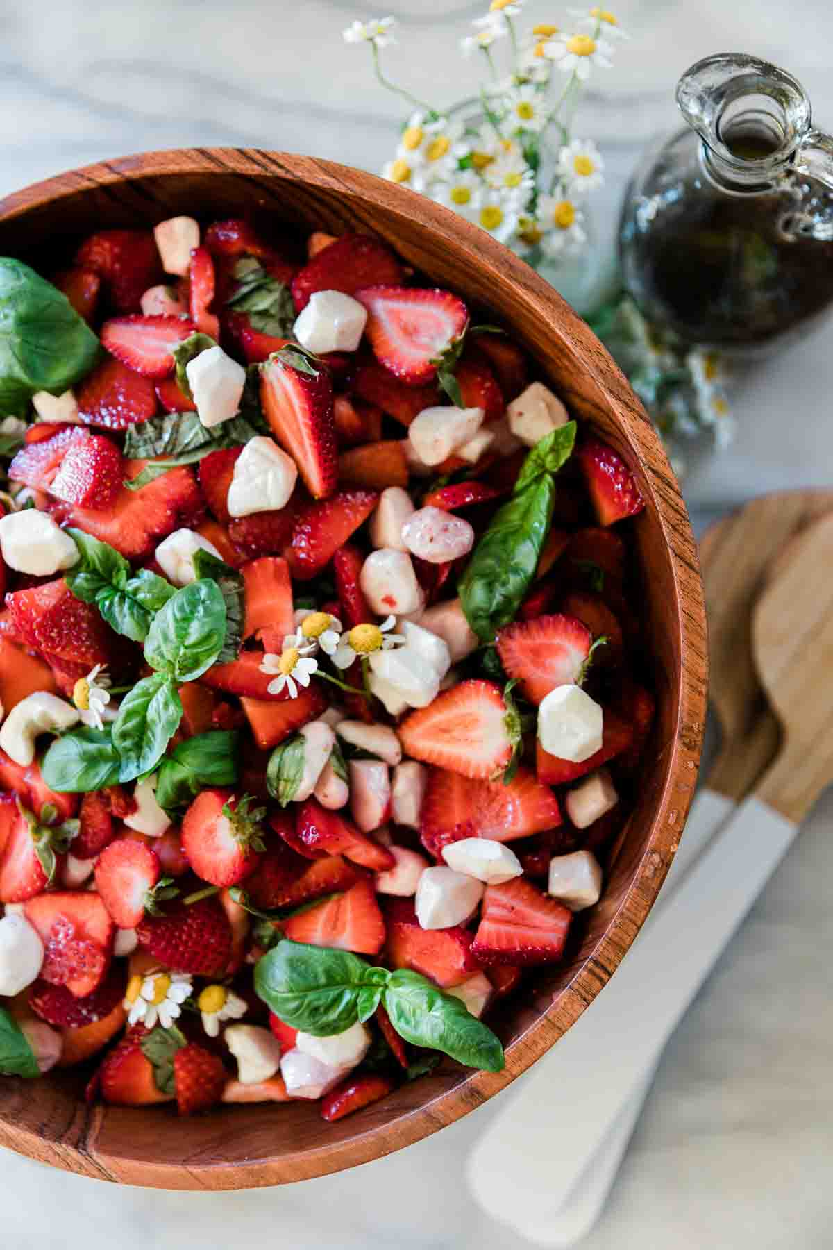 An overhead shot of strawberry salad in a wooden bowl. There are flowers to the side.
