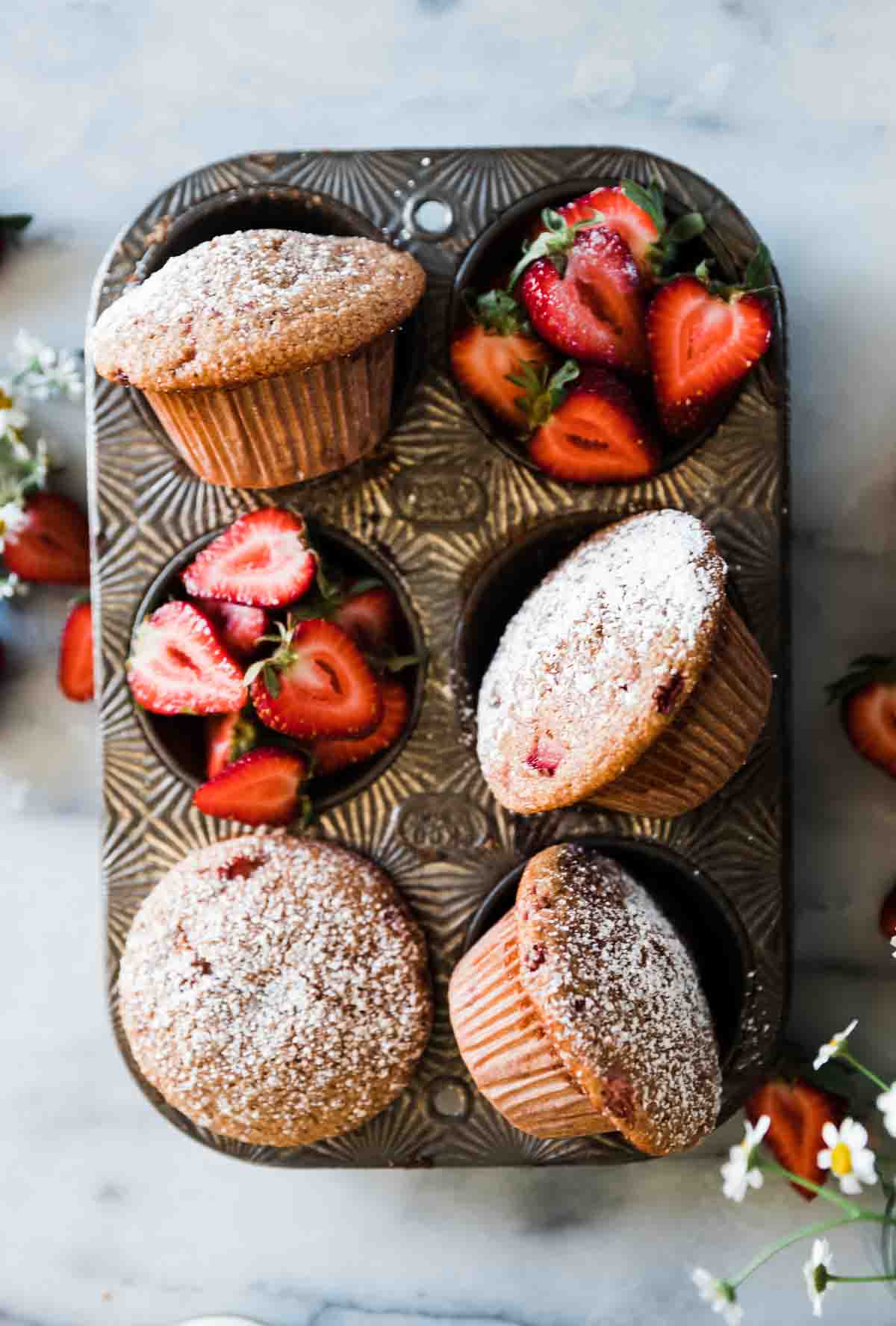 Healthy strawberry muffins in a muffin tin. There are two basins filled with strawberries.