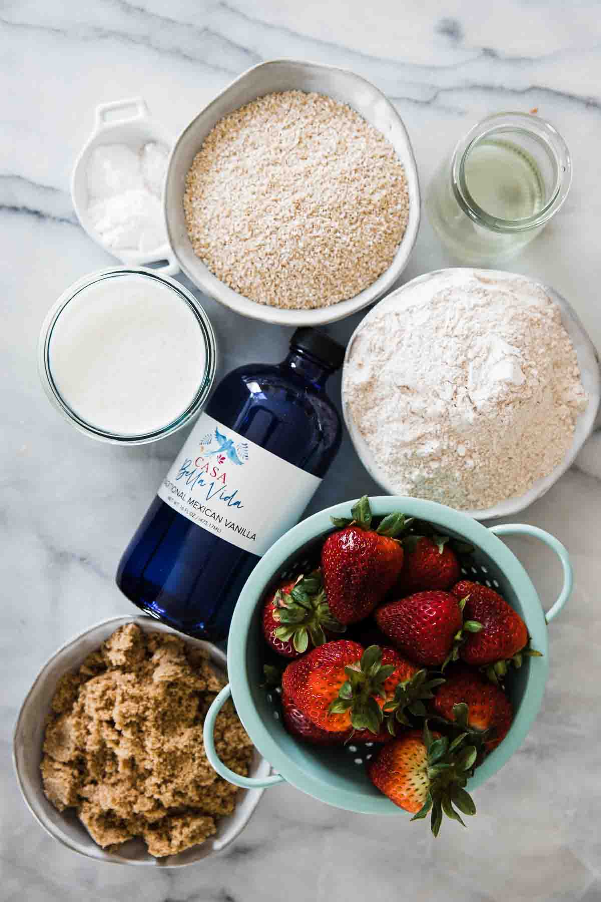 Oat bran, wheat flour, milk, vanilla, sugar and strawberries on a marble counter.