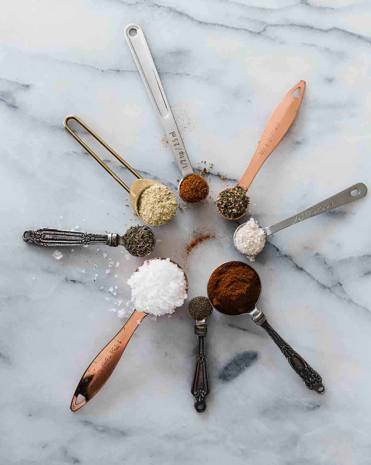 Spices in measuring spoons arranged in a circle on a marble counter.