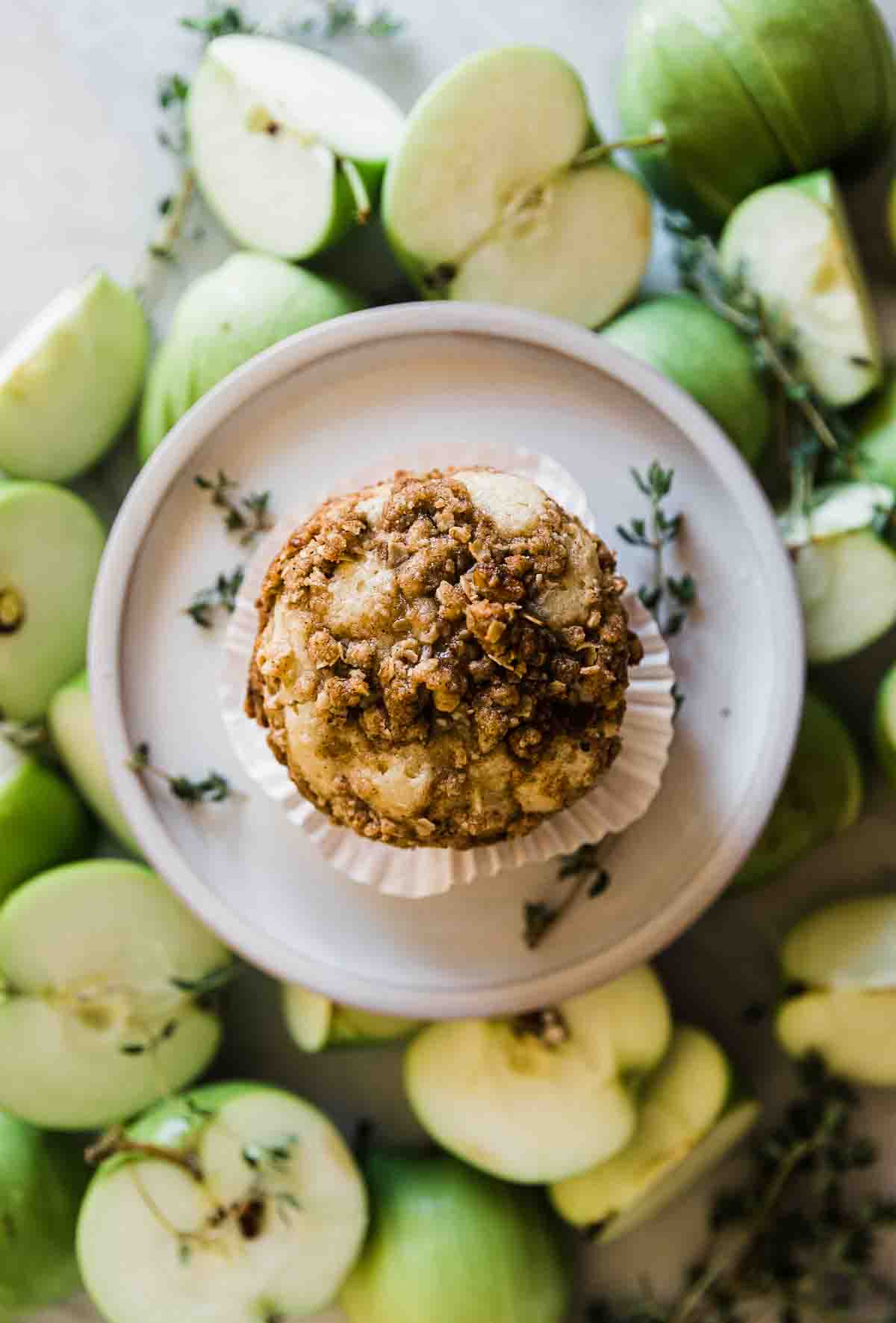 An overhead shot of apple muffin recipe. There is a single muffin on a plate surrounded by apples.