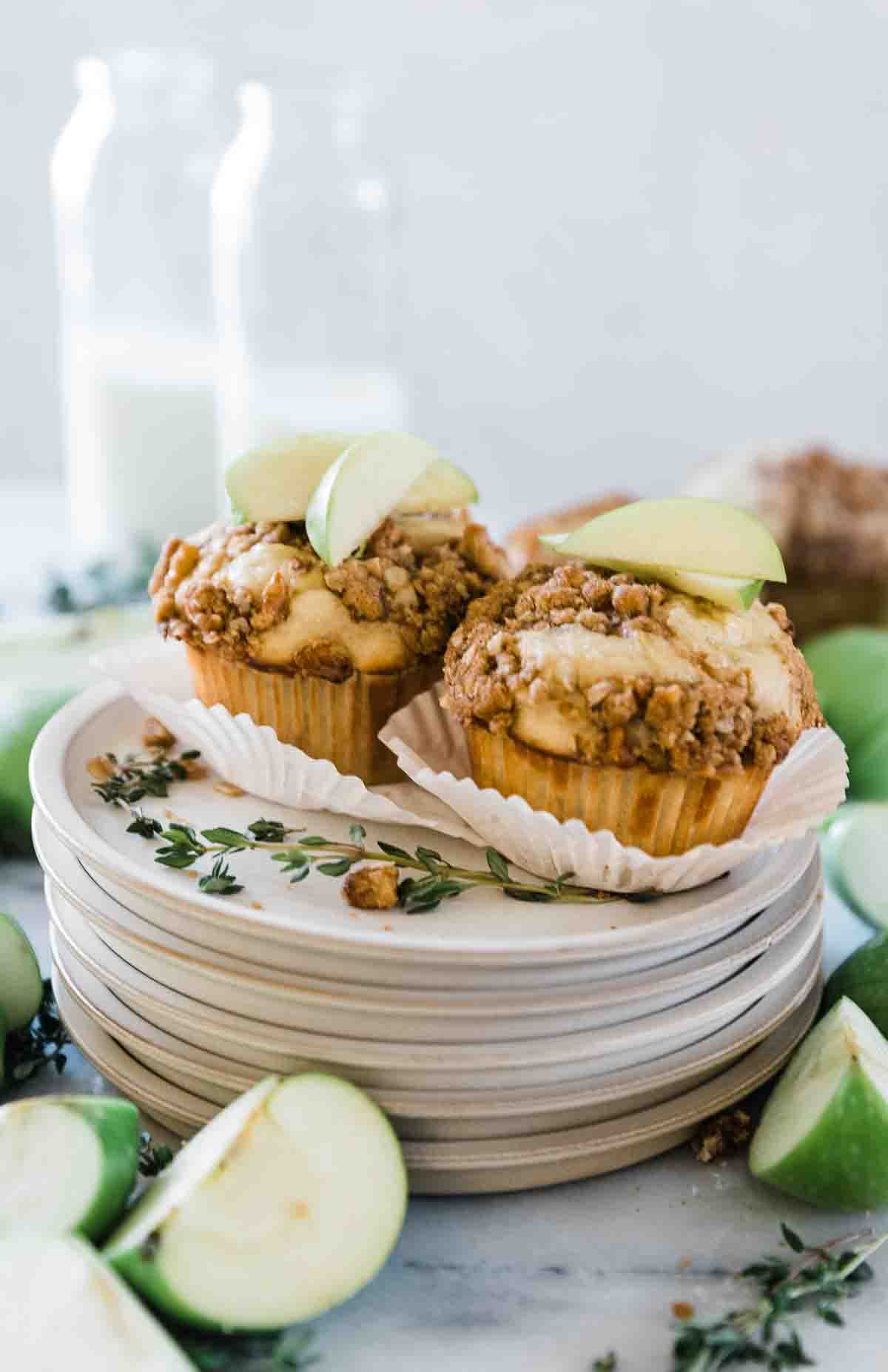 Two apple muffins on a stack of grey dessert plates. There are bottles of milk in the background.