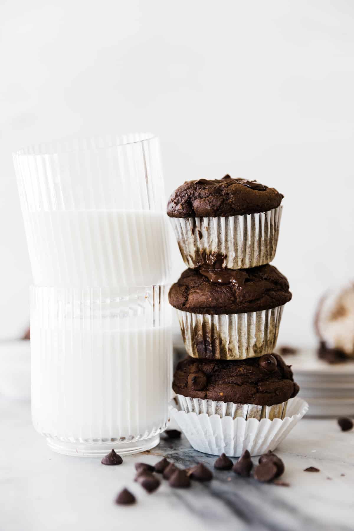 Three chocolate muffins stacked on top of each other next to a glass of milk. 