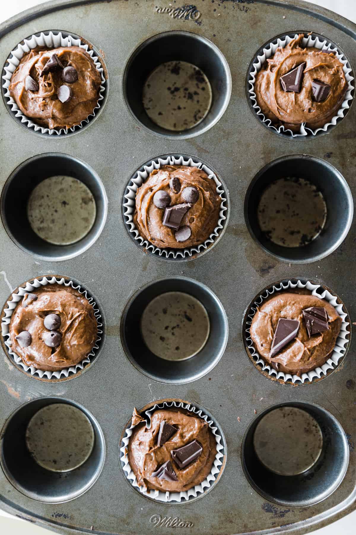 Batter in parchment lined muffin pan with chocolate chips on top of chocolate muffin batter. 