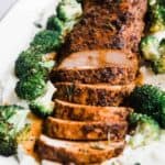 A close up of oven baked pork tenderloin on a white platter. It is on a bed of mashed potatoes and broccoli.