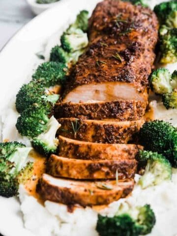 A close up of oven baked pork tenderloin on a white platter. It is on a bed of mashed potatoes and broccoli.