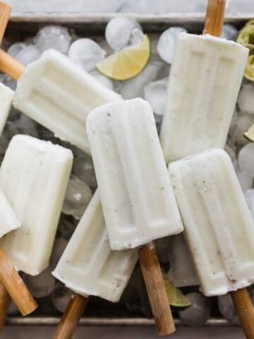 Creamy lime coconut popsicles on a tray with ice cubes and lime wedges.