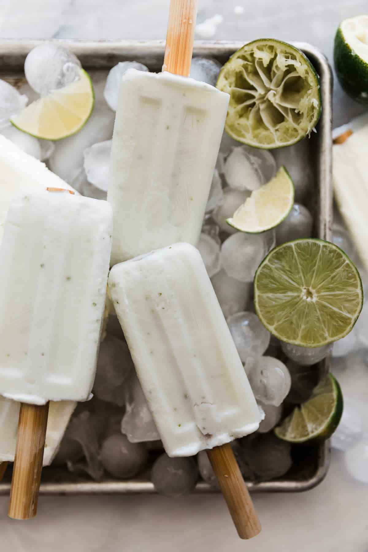 Three creamy lime popsicles on a tray of ice cubes and lime wedges. 