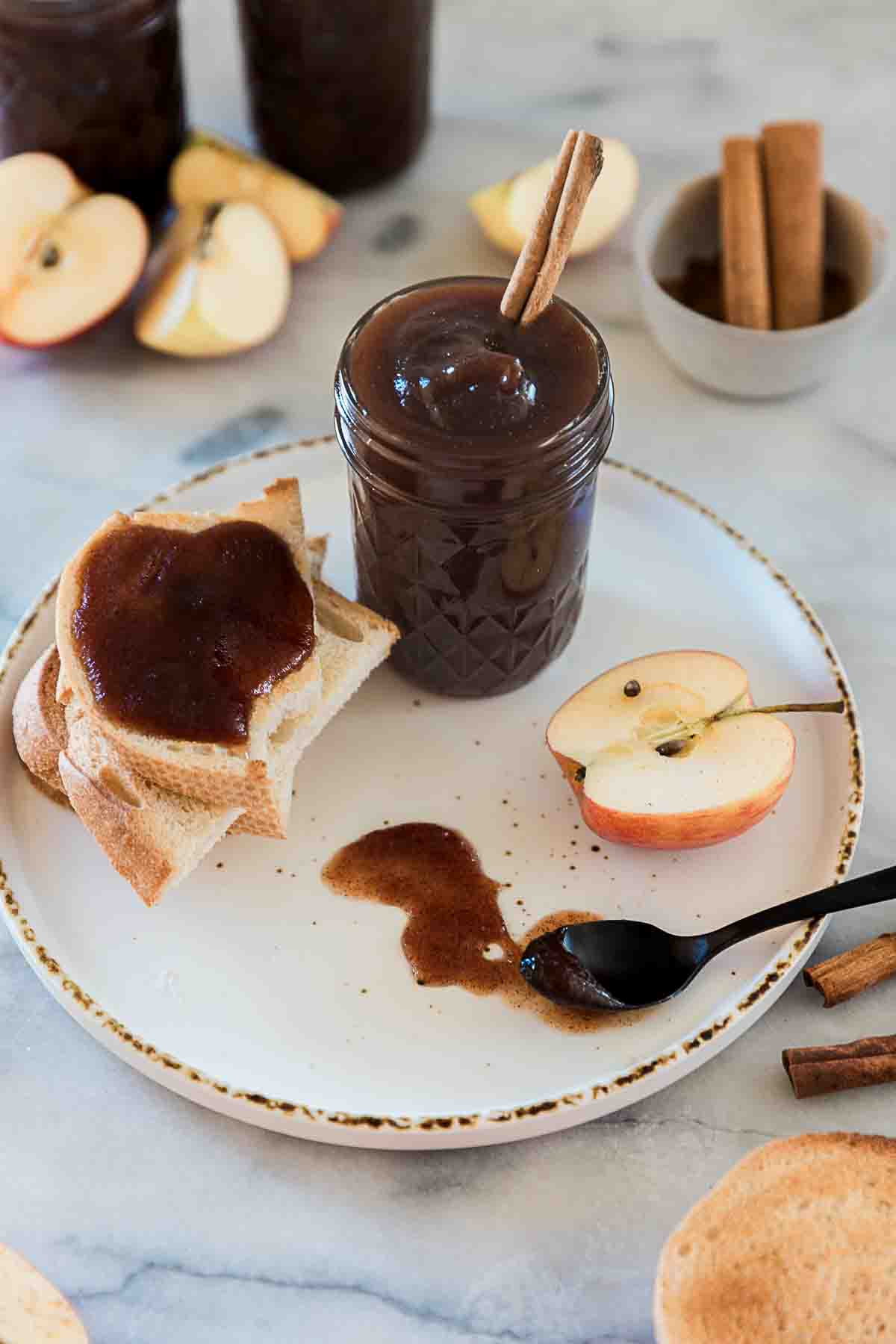 Apple butter in a mason jar. It is on a plate with bread and an apple.