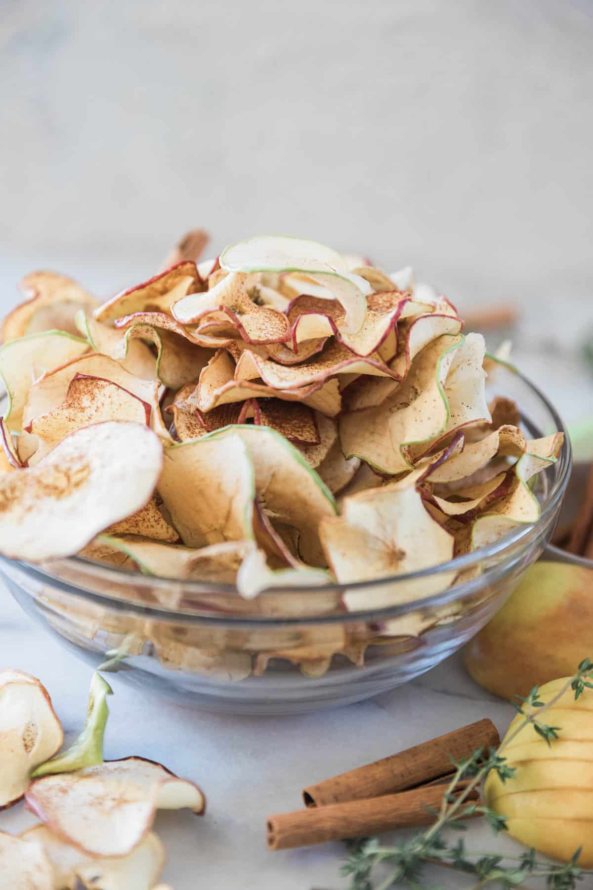 Apple chips in a glass bowl. They are spilling over onto a marble counter.