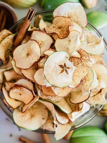 Apple chips in a glass bowl. There are sliced apples surrounding it.