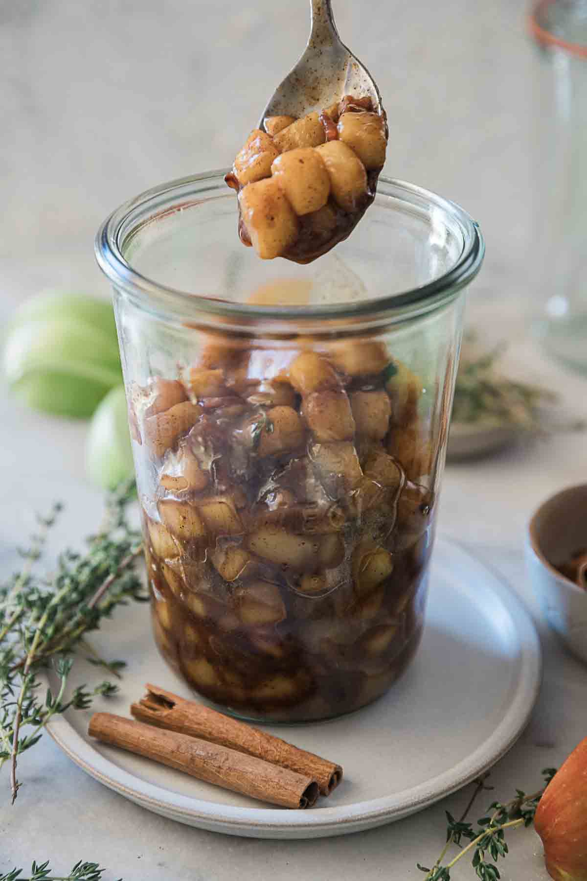 Cinnamon apples in a glass jar. There are cinnamon sticks and thyme to the side.