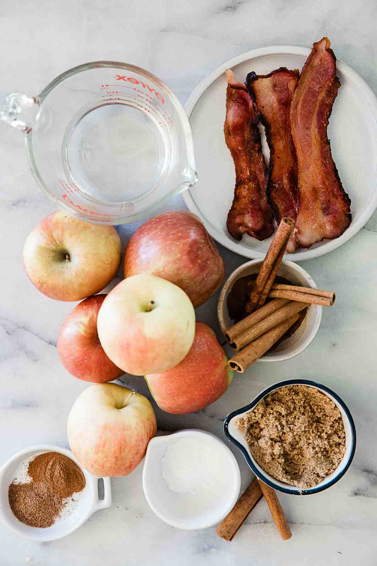Apples, cinnamon, brown sugar, and bacon on a marble counter.