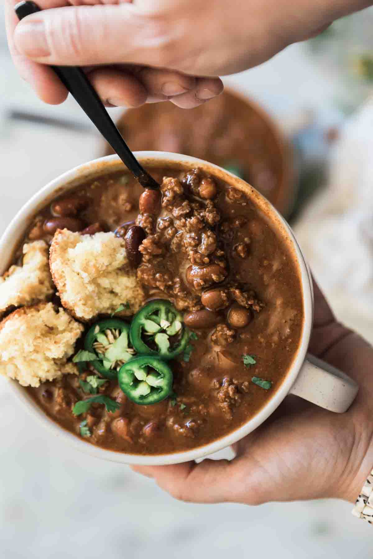 A bowl of chili being held with a spoon inserted. It is garnished with cornbread and jalapeños.
