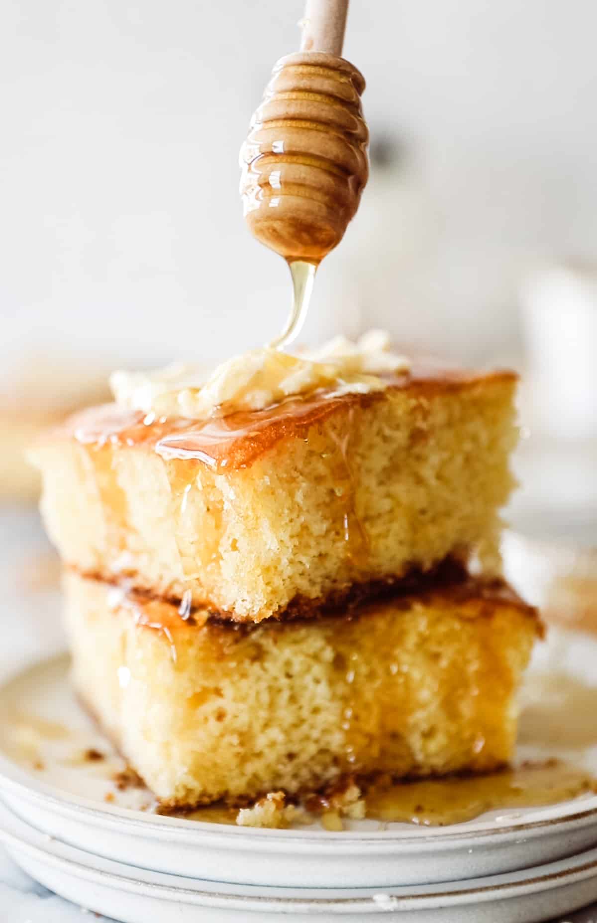 Two fluffy pieces of cornbread stacked on top of each other with butter and honey being drizzled over. 