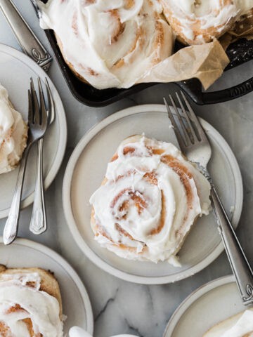 One cinnamon roll on a plate in the middle surrounded by a pan and other plates of cinnamon rolls.
