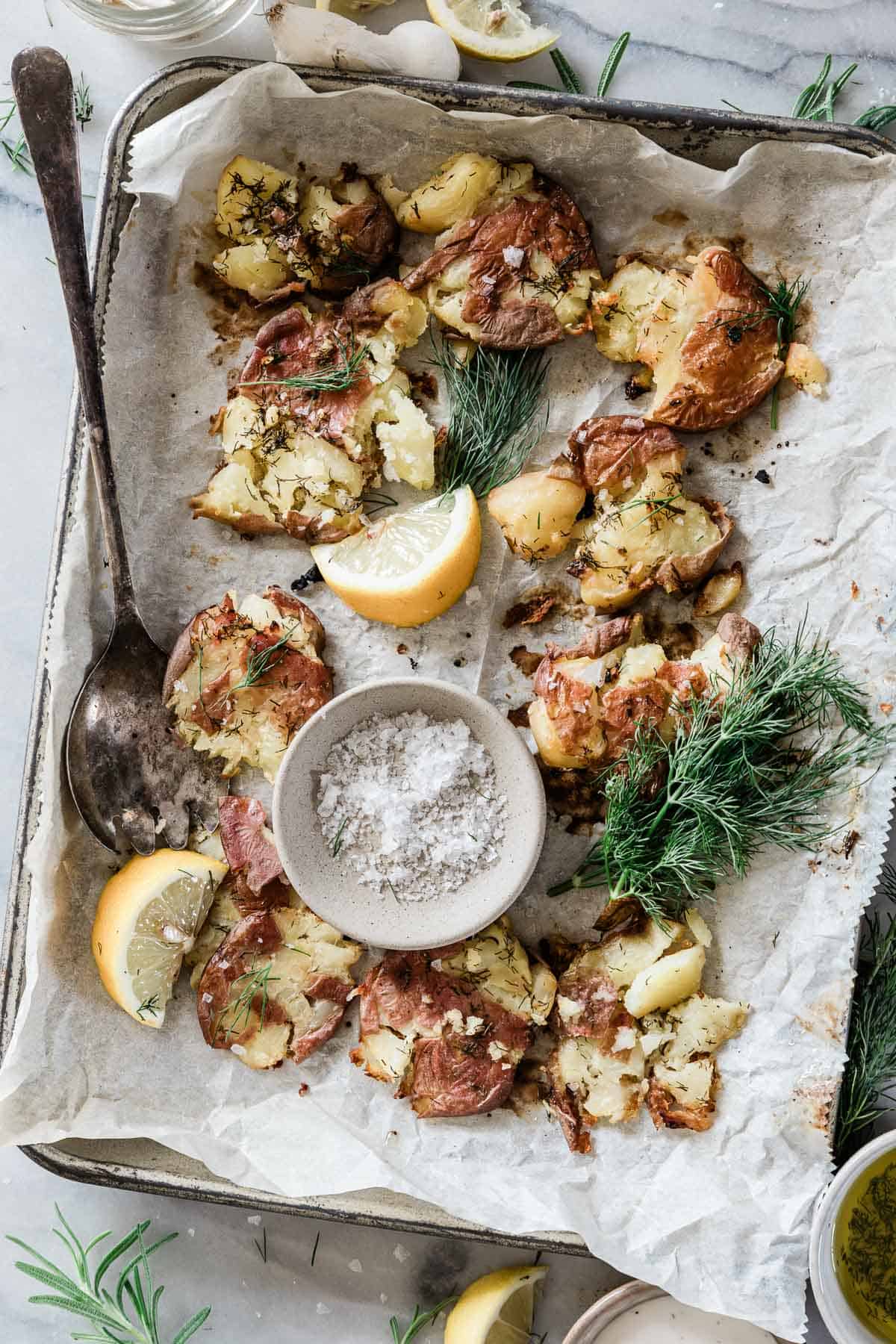 Smashed red potatoes on a baking tray lined with parchment. They are garnished with dill and lemon.