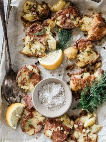 Smashed red potatoes on a baking tray lined with parchment. They are garnished with dill and lemon.
