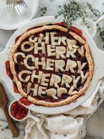 Homemade cherry pie in a white pie dish. There is thyme and cherries to the side.
