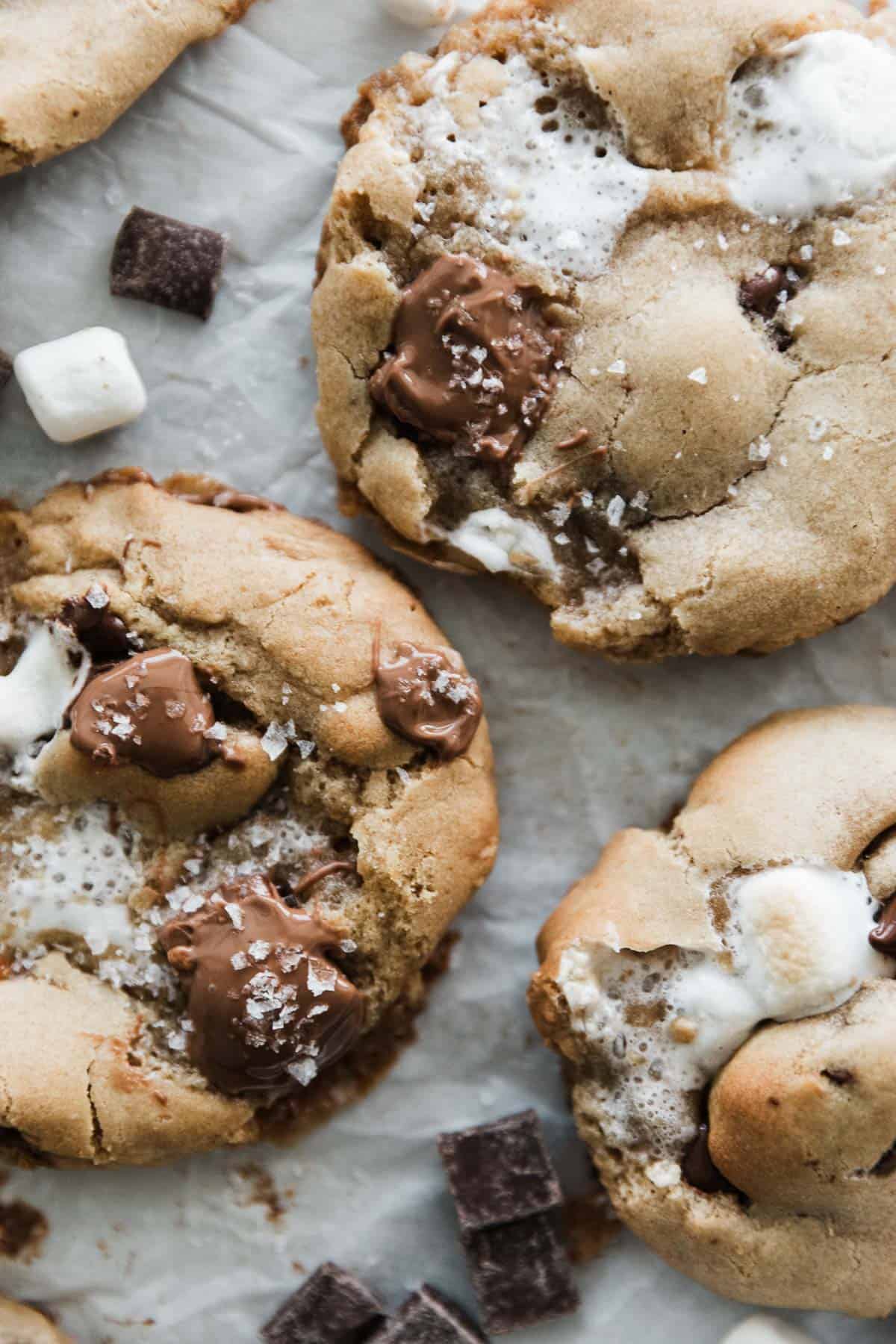 Three cookies with gooey marshmallows and chocolate pools on parchment paper with chocolate chips around them. 
