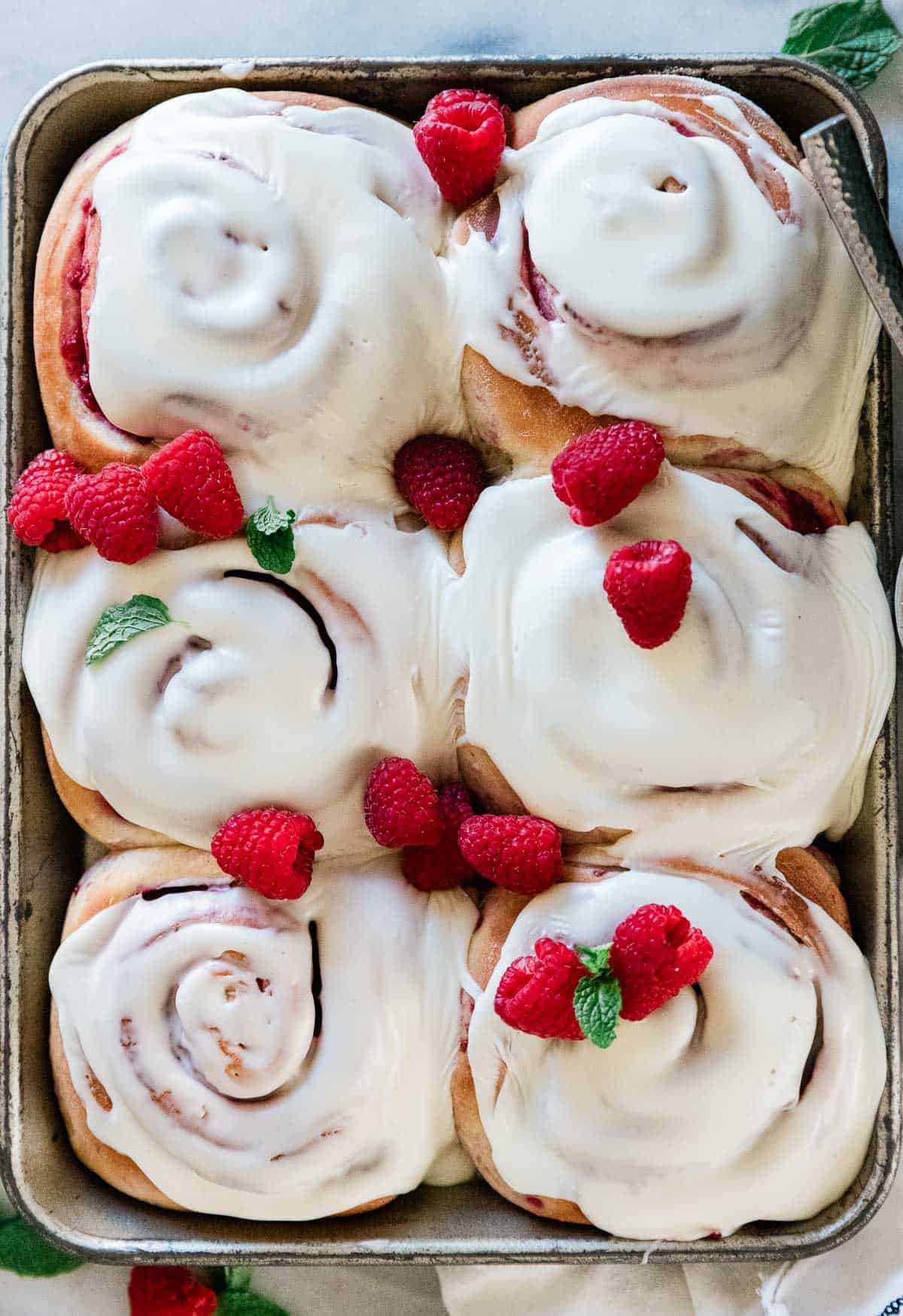 A pan of raspberry rolls. They are frosted and garnished with mint and raspberries.